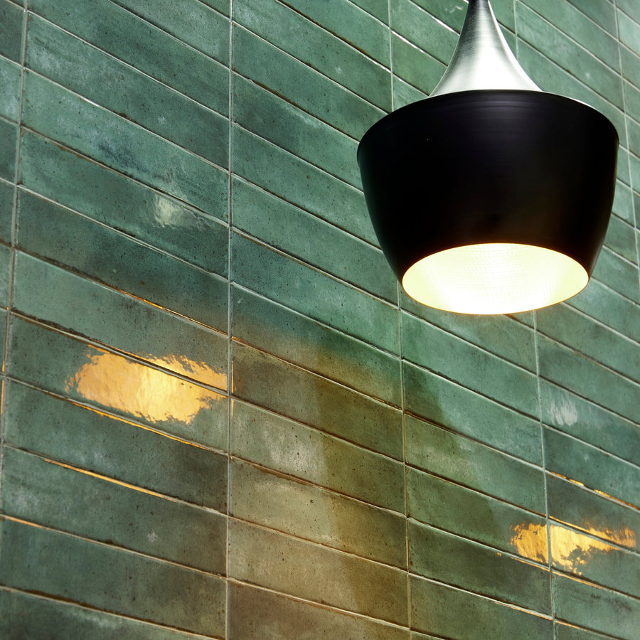 A modern black pendant light illuminates textured green wall tiles, highlighting subtle gold reflections and shadows.