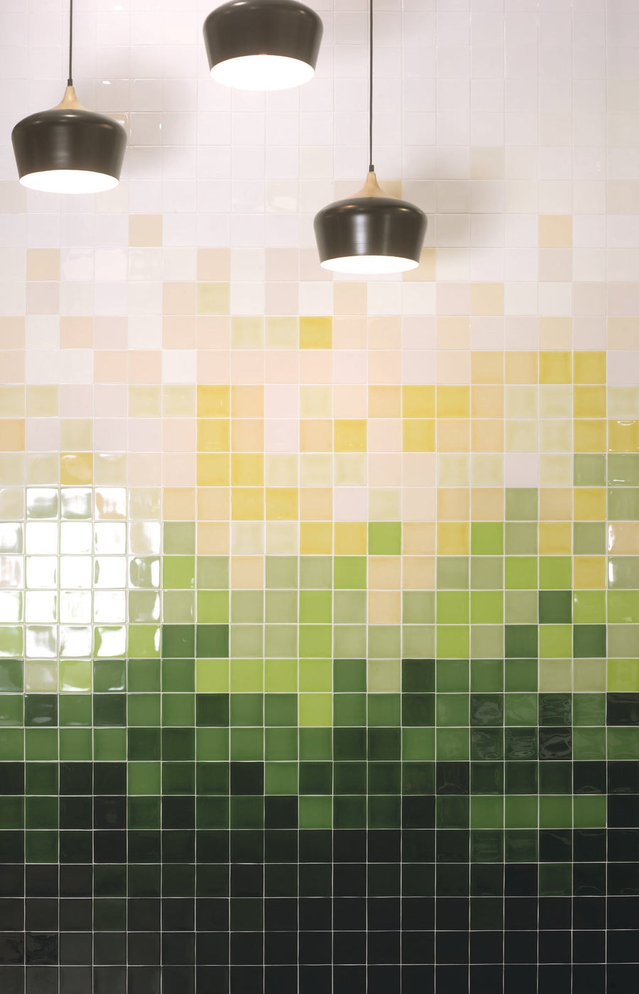 A vibrant mosaic wall featuring gradient shades of green and yellow tiles, complemented by three black pendant lights above.