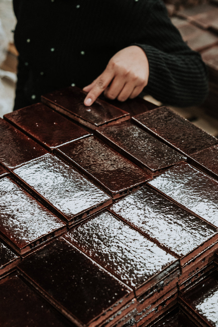 A hand wearing a black sleeve points at a stack of shiny, rectangular, warm brown colored tiles arranged neatly.