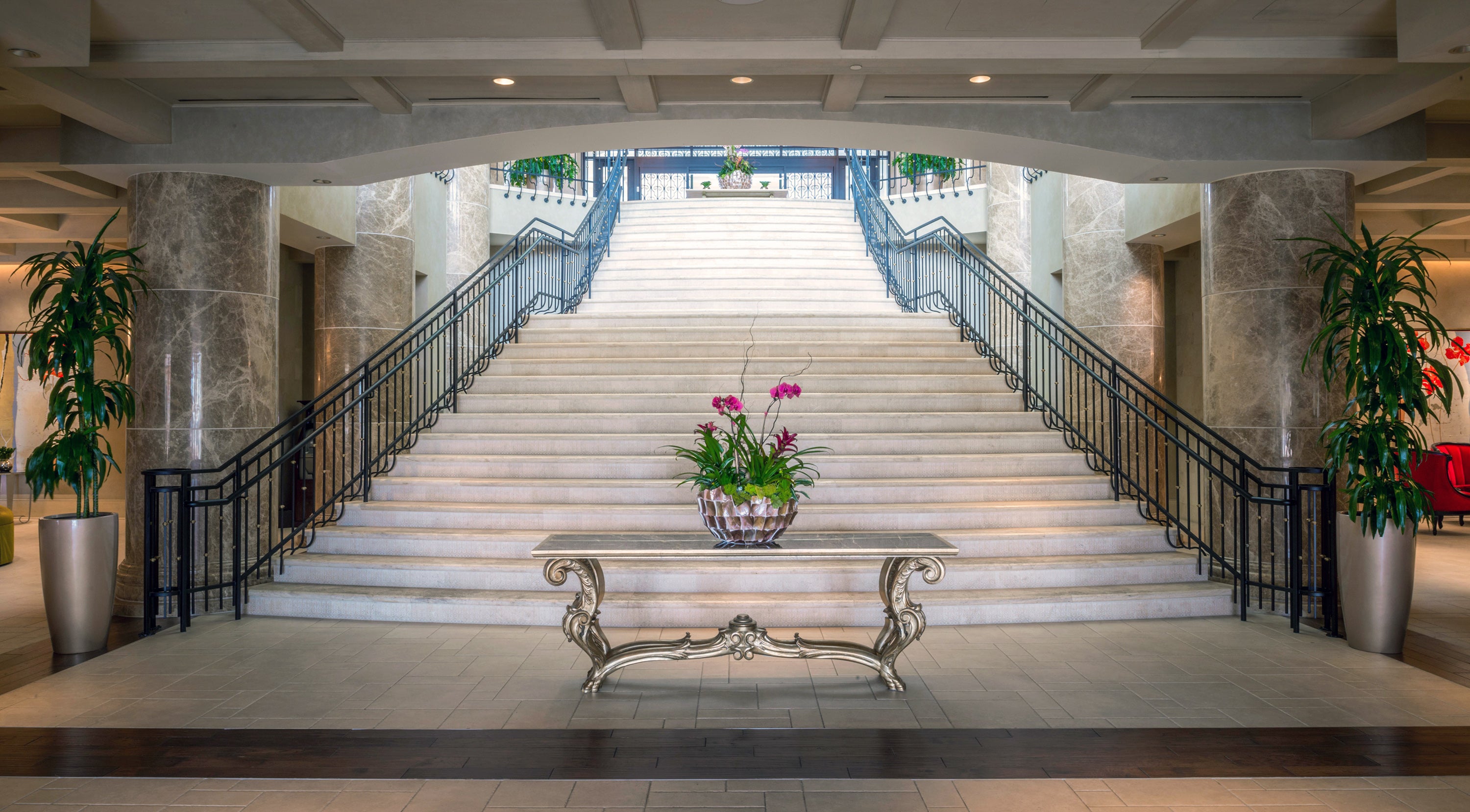 Elegant hotel lobby featuring a grand stone staircase, potted plants, and a decorative table with flowers at the center.