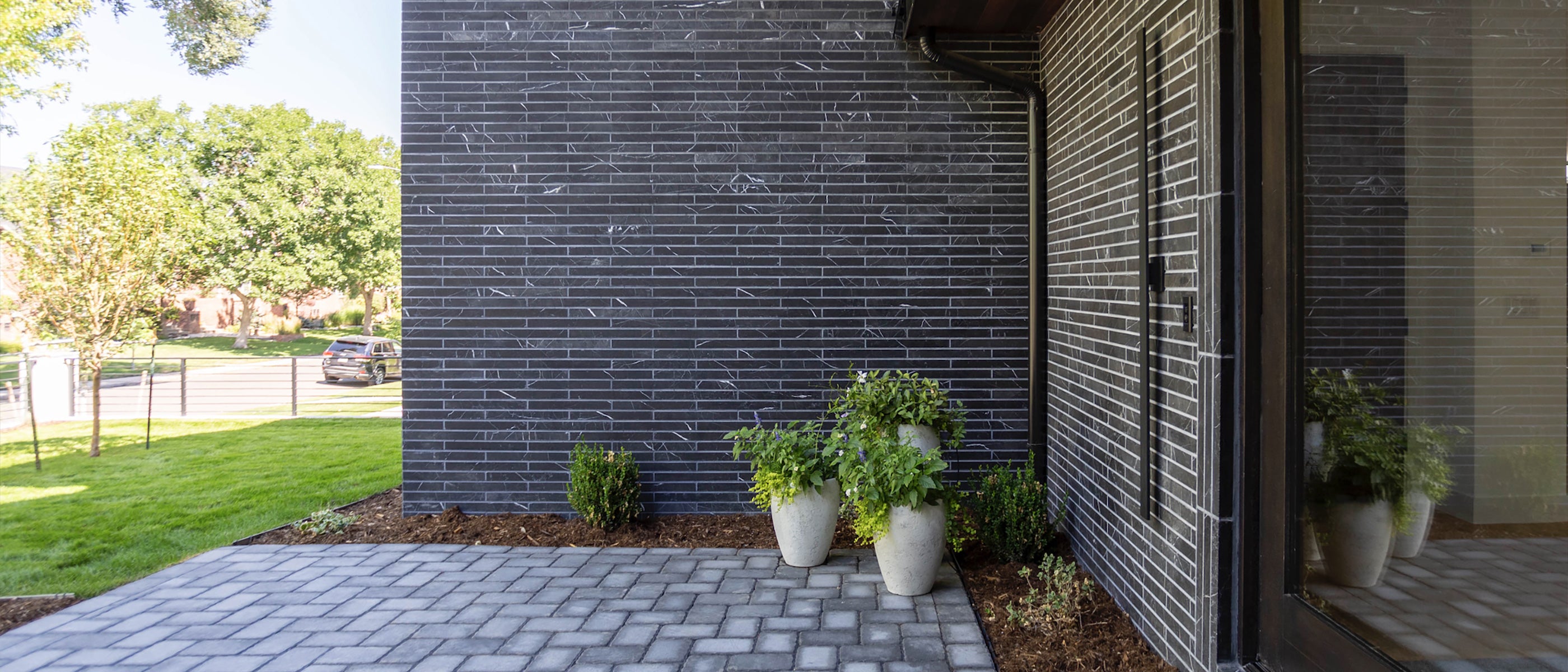 A modern entrance with a black brick wall, paver stones, and greenery in decorative pots, viewed from a sunny outdoor patio.