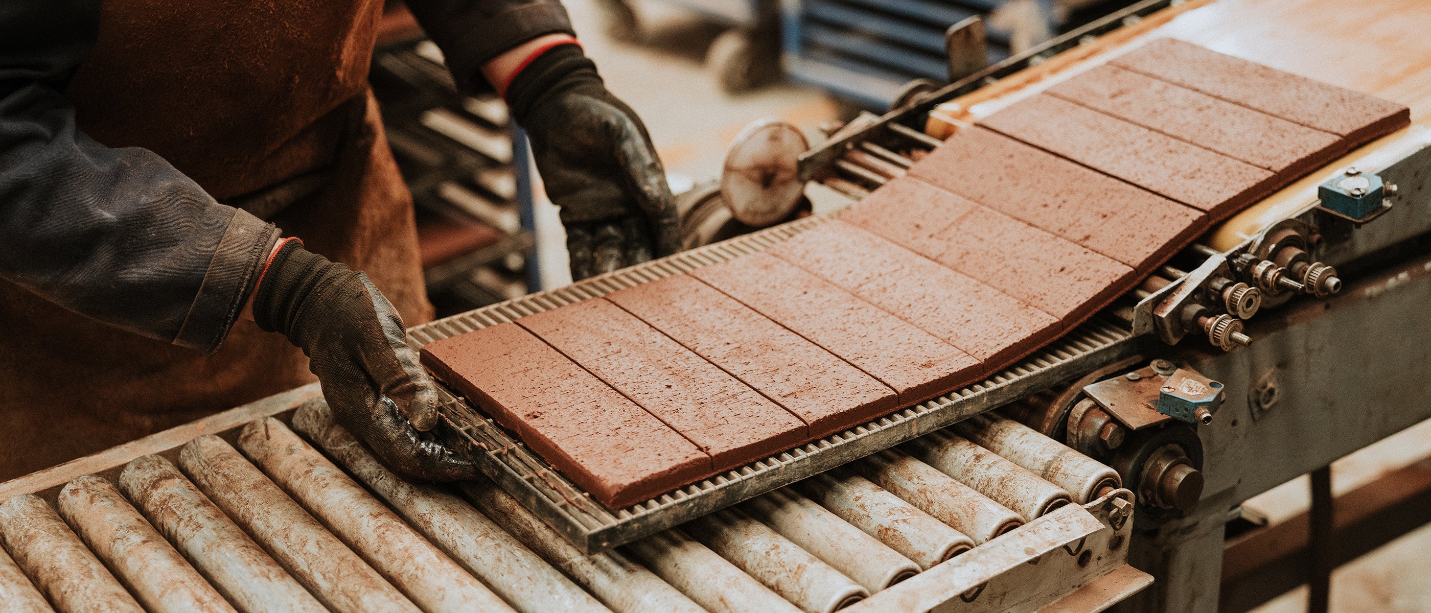 A worker in gloves handles freshly formed ceramic tiles on a conveyor belt in a factory setting.