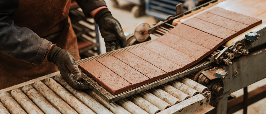 A worker in gloves handles freshly formed ceramic tiles on a conveyor belt in a factory setting.